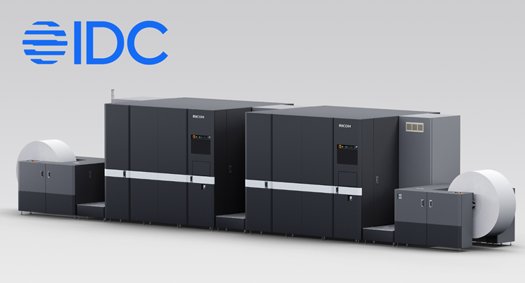  large industrial printing machine with two main black units connected in series, each featuring control panels and silver horizontal stripes. The setup includes large white paper rolls on both ends, indicating continuous paper feed for high-volume printing, with IDC logo in blue on the top left corner.